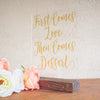 Clear acrylic dessert sign that reads "first comes love, then comes dessert" in gold writing on a wood surface.